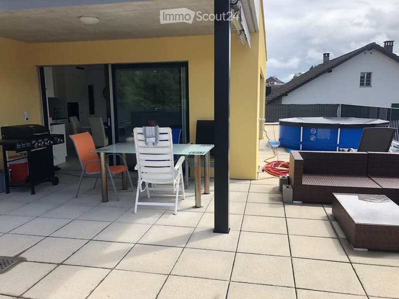 A covered patio area with tiled flooring, featuring a glass table with chairs, a wicker sofa set, and a barbecue grill. A small inflatable pool is visible in the background.