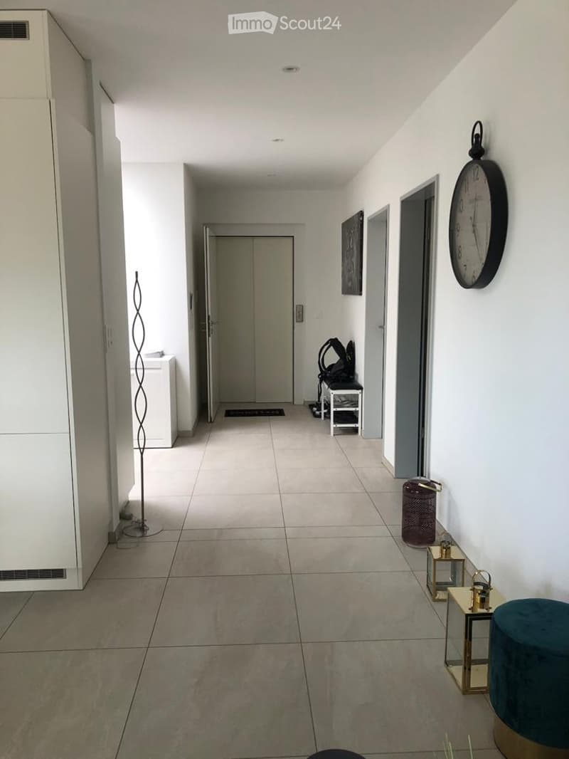 Clean, modern hallway with tiled flooring, white walls, and several doors leading to different rooms. A tall, slender floor lamp stands on the left, and a small table with a blue cushioned stool is on the right. A wall-mounted clock and a small decorative