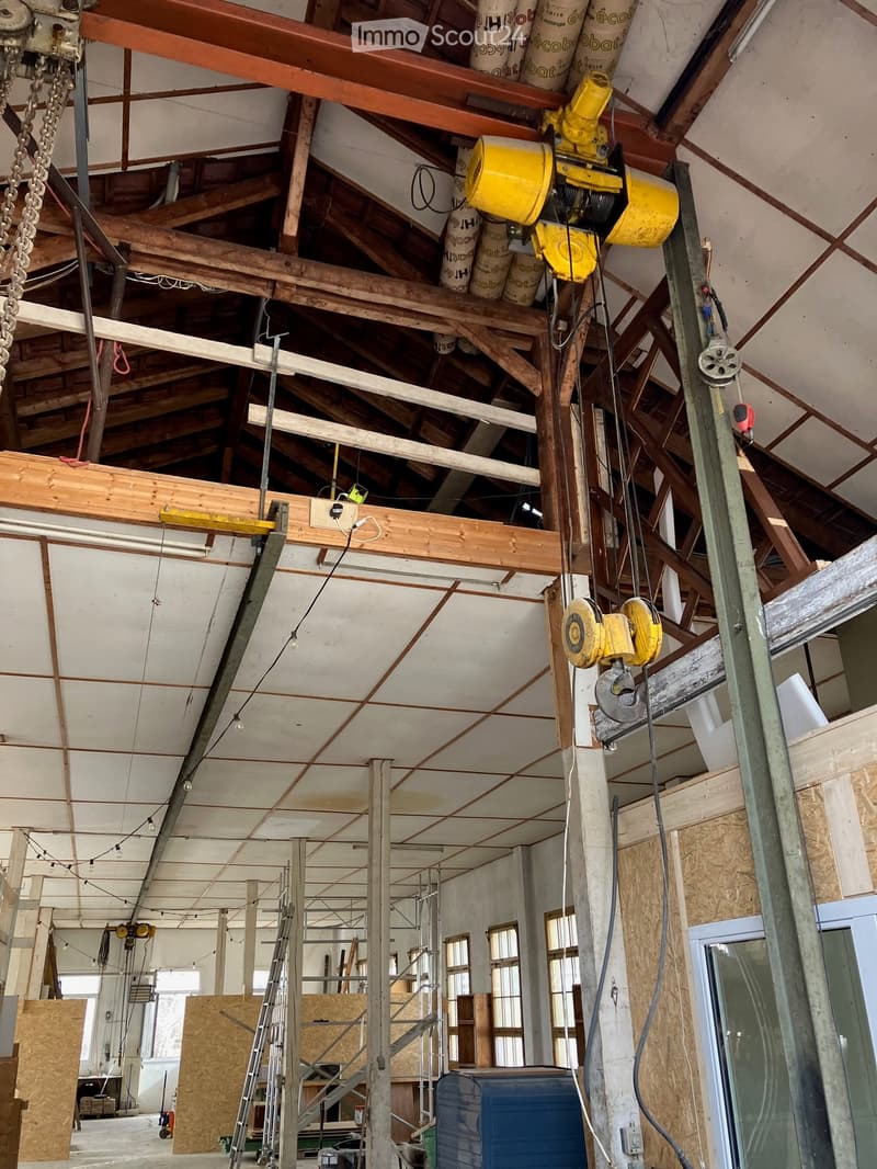An unfinished room with exposed wooden beams, metal scaffolding, and construction materials. The ceiling features exposed wooden beams and ductwork, with visible electrical wiring and a yellow industrial crane hanging from the ceiling.