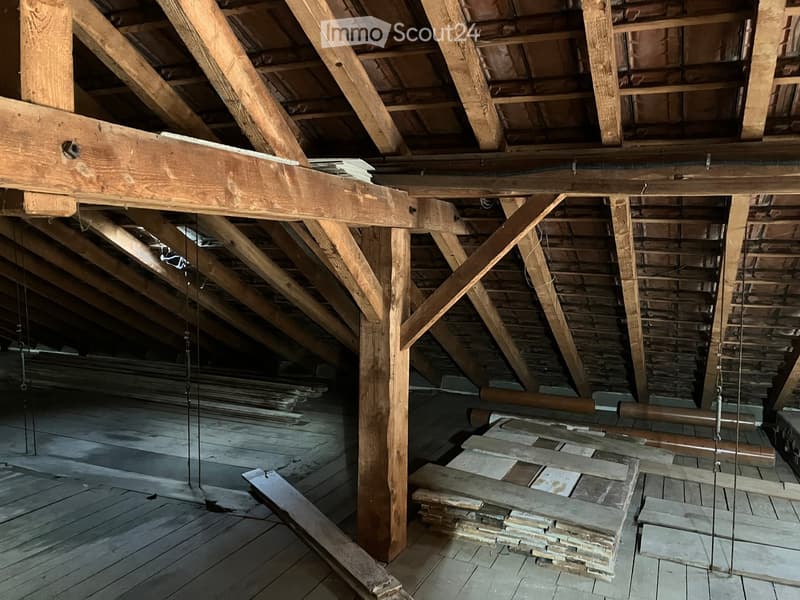 An attic space with exposed wooden beams and trusses, wooden floor, and stacks of wooden planks and boards stored on the floor.