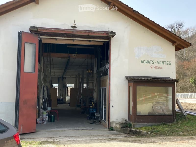 Two-story building with a large open garage door, visible tools and materials inside, and a storefront with signage reading 'ACHATS VENTES St. Blaise'.