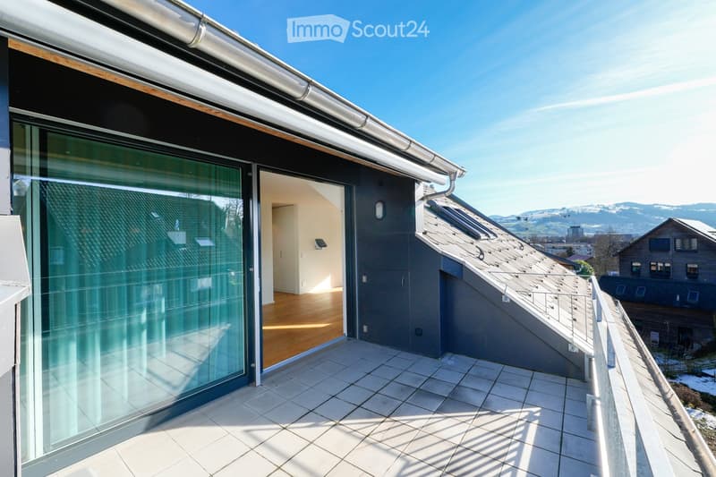 Modern balcony with tiled flooring, glass sliding doors leading to an interior space, and a view of a sloped roof and distant landscape.