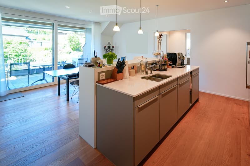 Modern kitchen with white countertops, wooden cabinets, and a central island. Features a sink, coffee maker, and a small dining area with a table and chairs. The space is well-lit with pendant lights and has a view of a balcony through large glass doors.