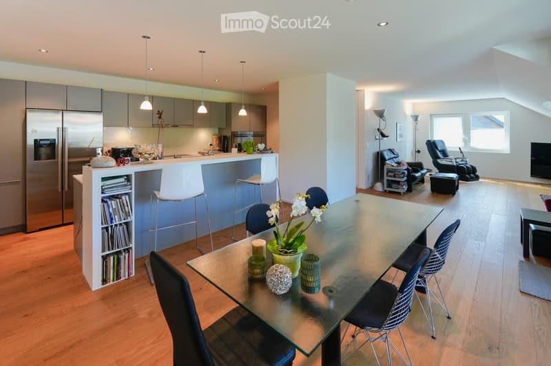 Modern open-plan living and dining area with a sleek kitchen, bar counter, and comfortable seating. The space features a glass dining table with chairs, a bar area with stools, and a living area with a TV and recliners.