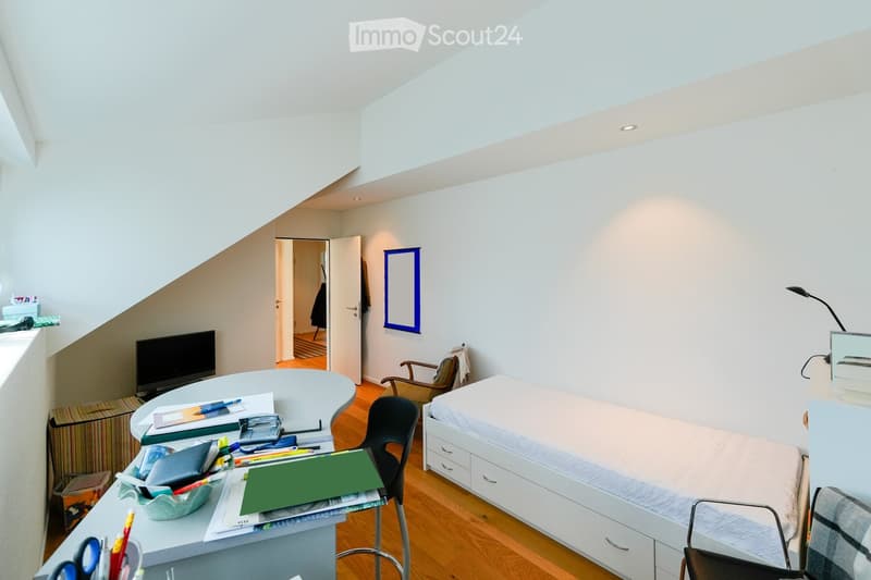 A modern, well-lit room with a bed, desk, and chair. The room features wooden flooring, white walls, and a slanted ceiling with skylights. A desk is cluttered with office supplies, and a bed with drawers is positioned against the wall.