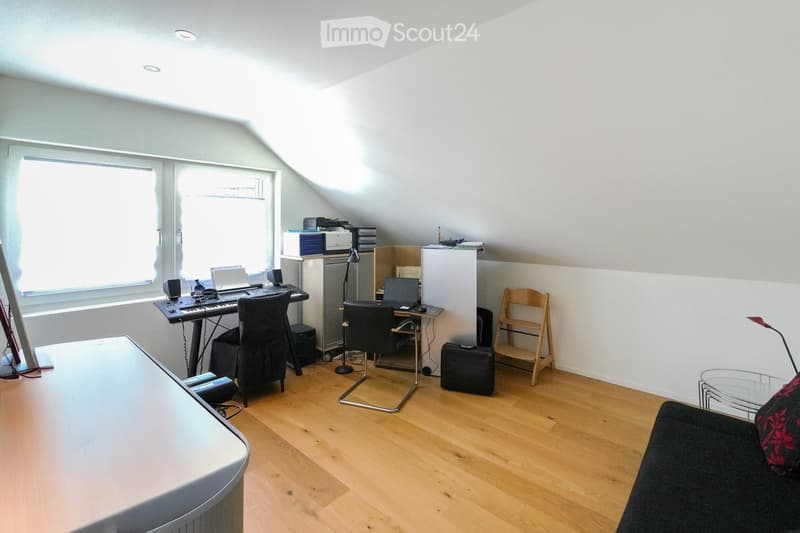 A multi-functional room with wooden flooring, a slanted ceiling, and large windows. Contains a sofa, a chair, a table with a laptop and keyboard, a printer, a refrigerator, a lamp, and a stool.