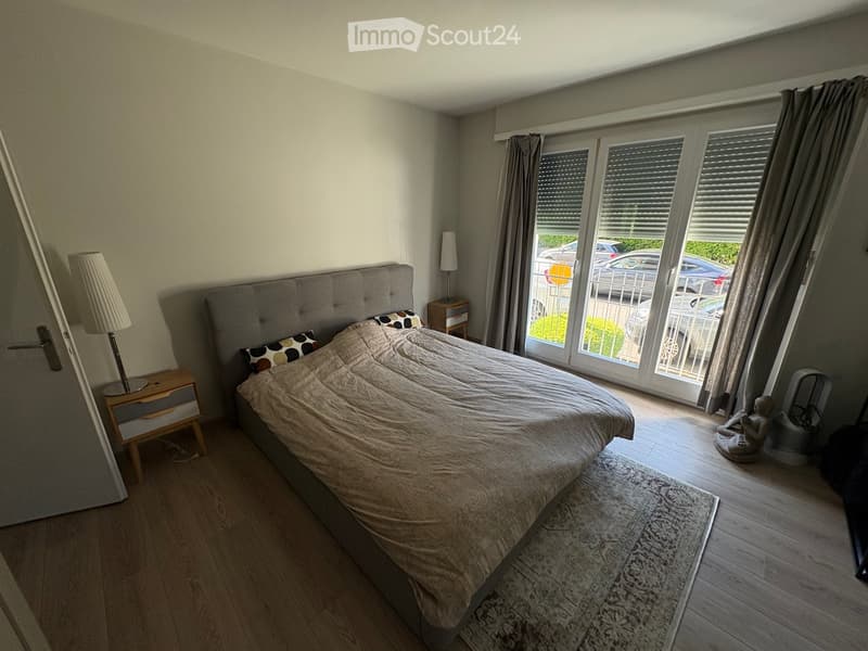 Well-lit bedroom with wooden flooring, large bed with grey headboard and beige bedding, nightstands with lamps, and large windows with curtains leading to a balcony.