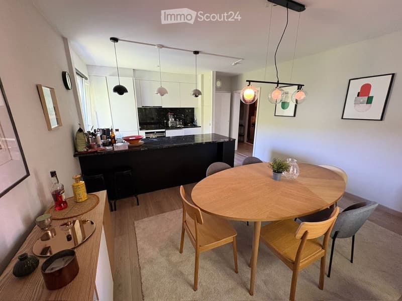 Open-plan living and dining area with a round wooden dining table, chairs, a kitchen island, pendant lights, and decorative items on a sideboard.