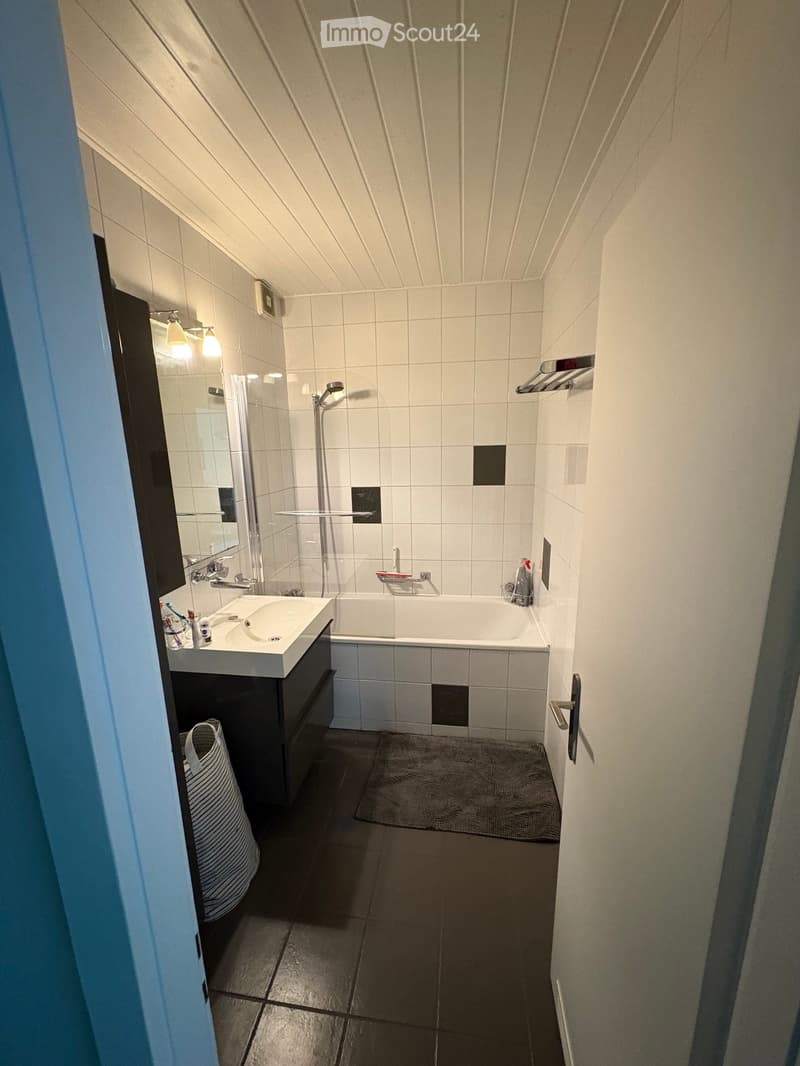 Modern bathroom with white tiles, a sink, bathtub, mirror, and shower stall. Features a dark grey floor and a small towel on the floor.