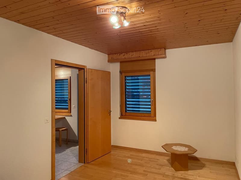Empty room with wooden flooring, white walls, and a wooden ceiling. Features a small table, a chair, and an open doorway leading to another room.