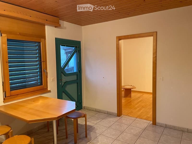 A small, unfurnished room with a wooden table and two stools, a green door, and a window with wooden shutters. The floor is tiled, and the walls are painted white.