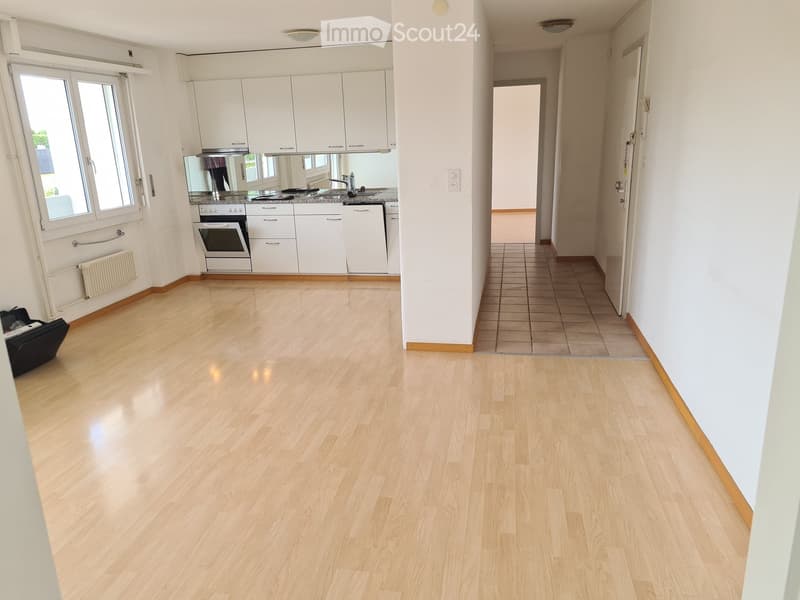 Empty hallway with light wooden flooring, white walls, and tiled section leading to another room. The hallway has a door at the end and a small section of a kitchen visible on the left side.