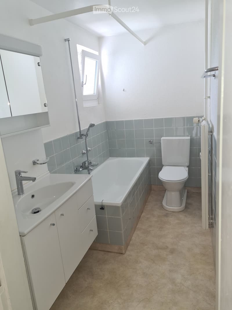 Modern bathroom with white walls, grey tiles, a bathtub, toilet, sink, and a window for natural light.