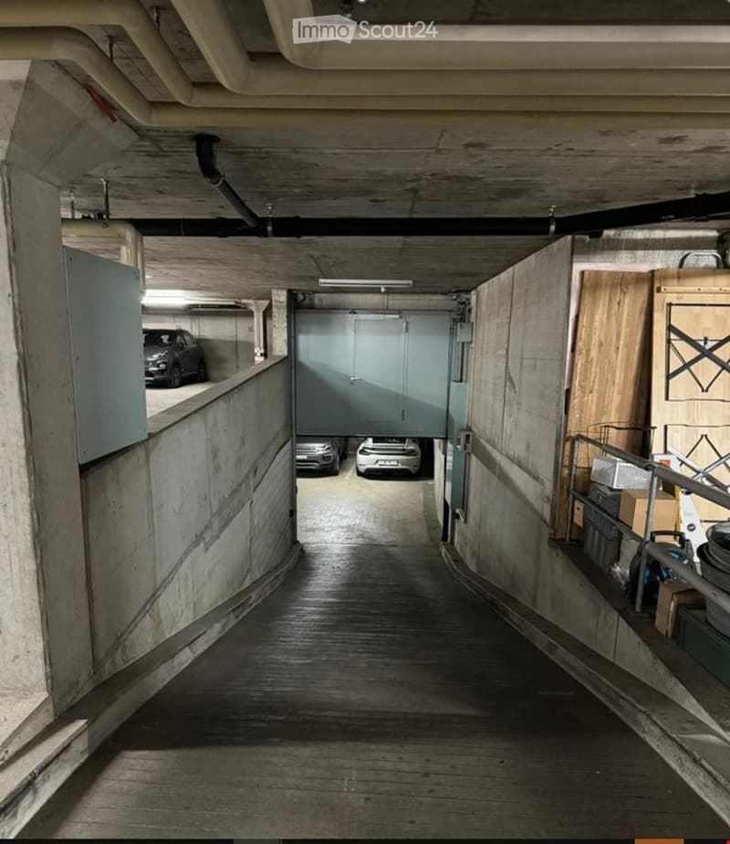A narrow, industrial-style corridor with exposed wooden beams and concrete walls, leading to a parking area with several cars visible in the background.