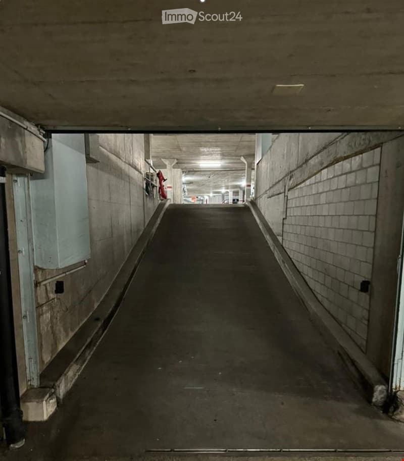 A dimly lit underground corridor with concrete walls and a smooth concrete floor, leading to a parking area in the distance.