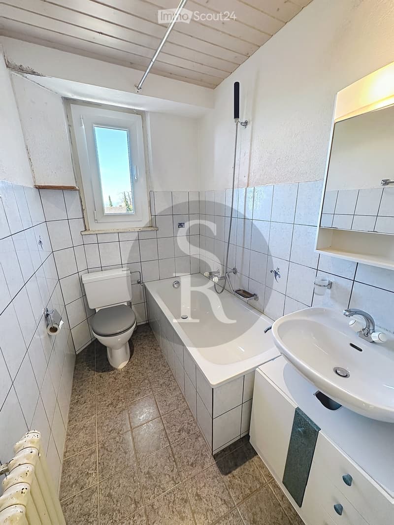Bathroom with a toilet, bathtub, sink, mirror, and cabinet. The room has tiled walls and a window letting in natural light.