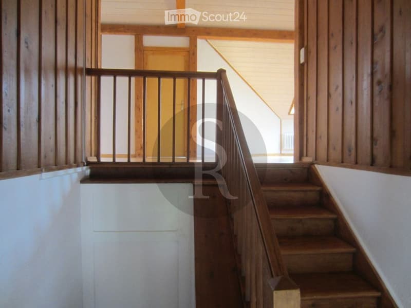 Wooden staircase with a handrail and open wooden balustrade, leading to an upper level with exposed wooden beams and white walls.
