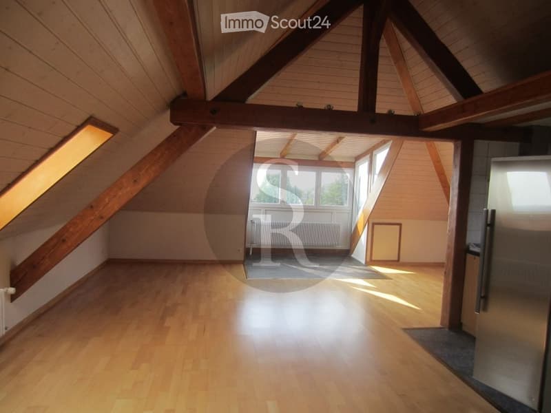 Bright, empty attic room with wooden floors and exposed wooden beams. Large windows allow natural light to fill the space. The room features a simple layout with a radiator and a small entryway leading to another room.