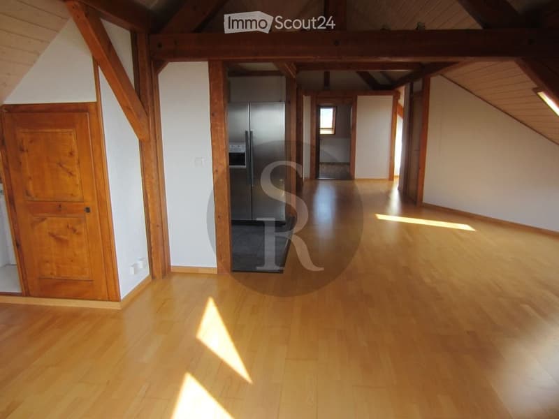 Empty room with wooden flooring, white walls, and exposed wooden beams. A stainless steel refrigerator is visible in the background, but the room itself is unfurnished.