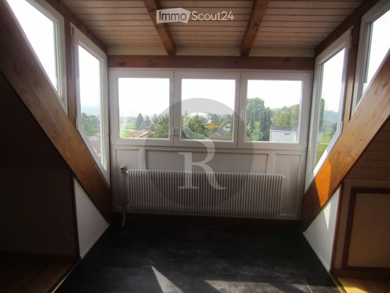 Empty room with wooden ceiling beams, large windows offering a scenic view, radiator, and dark flooring.