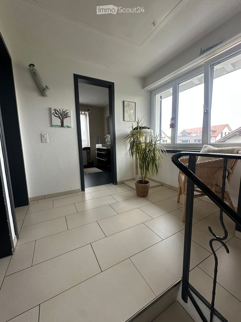 Bright balcony with tiled floor, large windows offering a view, a potted plant, and a chair near the railing.