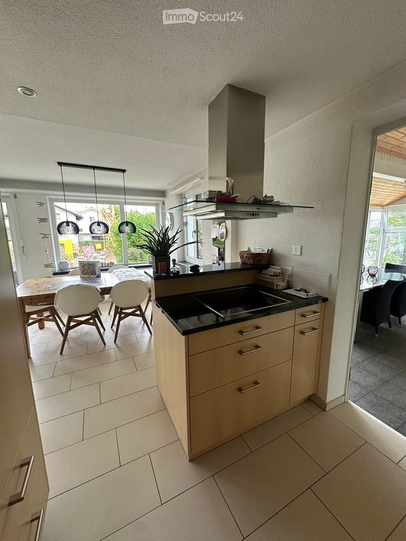 Modern kitchen with light-colored cabinets, black countertop, induction stove, range hood, pendant lights, and a small dining area with chairs and a table.