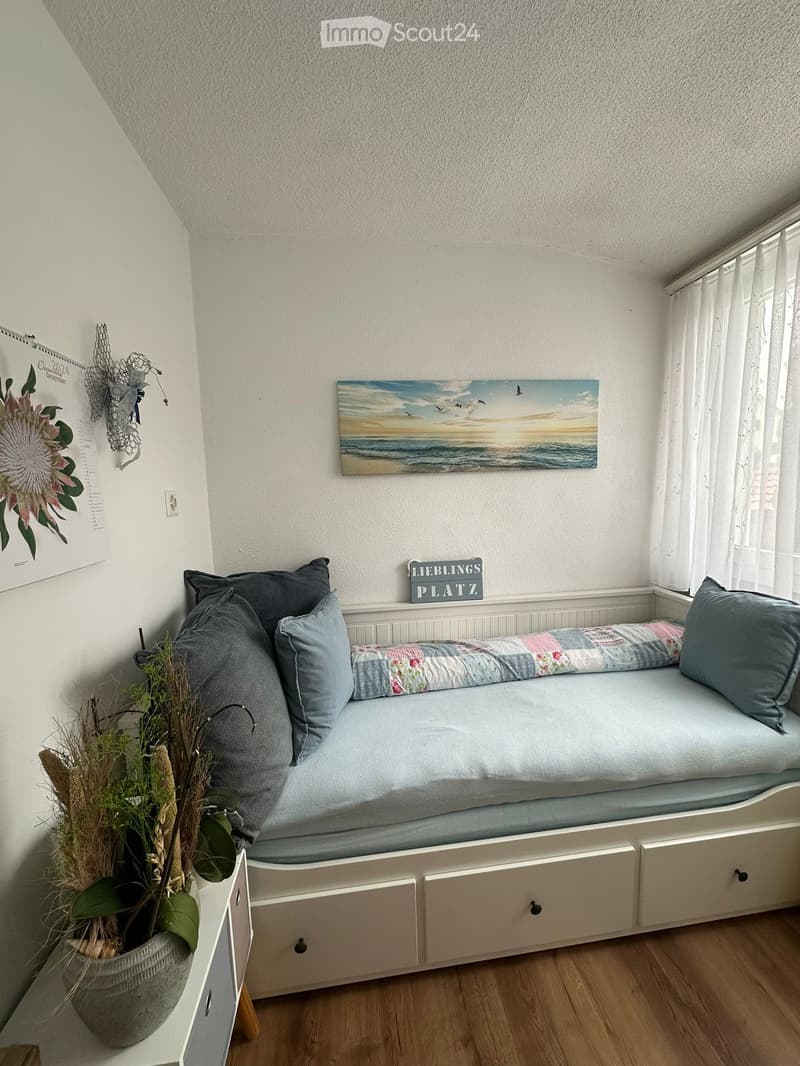 Cozy bedroom with a daybed, pillows, a nightstand with a vase, a painting, and a calendar on the wall.