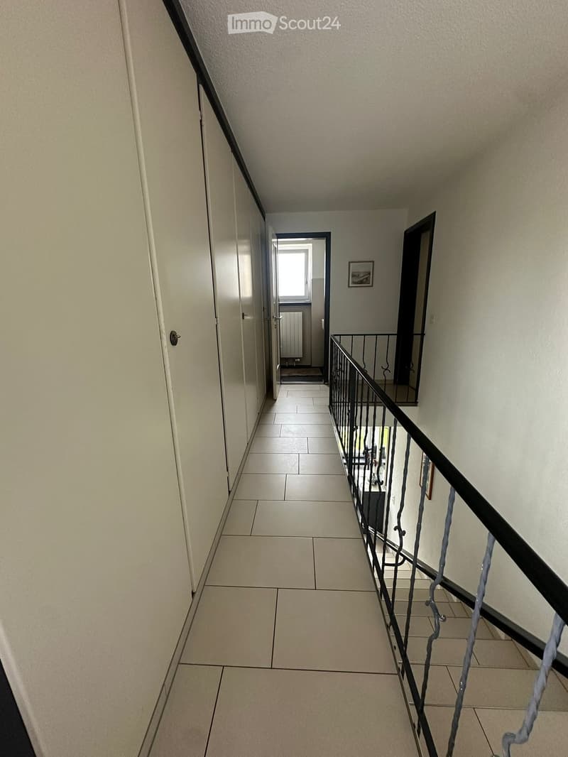 A narrow hallway with tiled flooring, white walls, and a black metal railing leading to an open area with a window and a radiator.