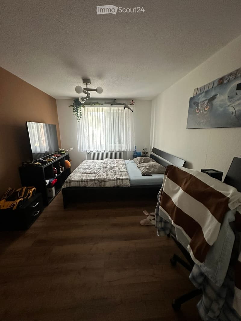 Cozy bedroom with a double bed, nightstand, mirror, chair, shelf, and a painting on the wall. The room features wooden flooring and a window with white curtains.