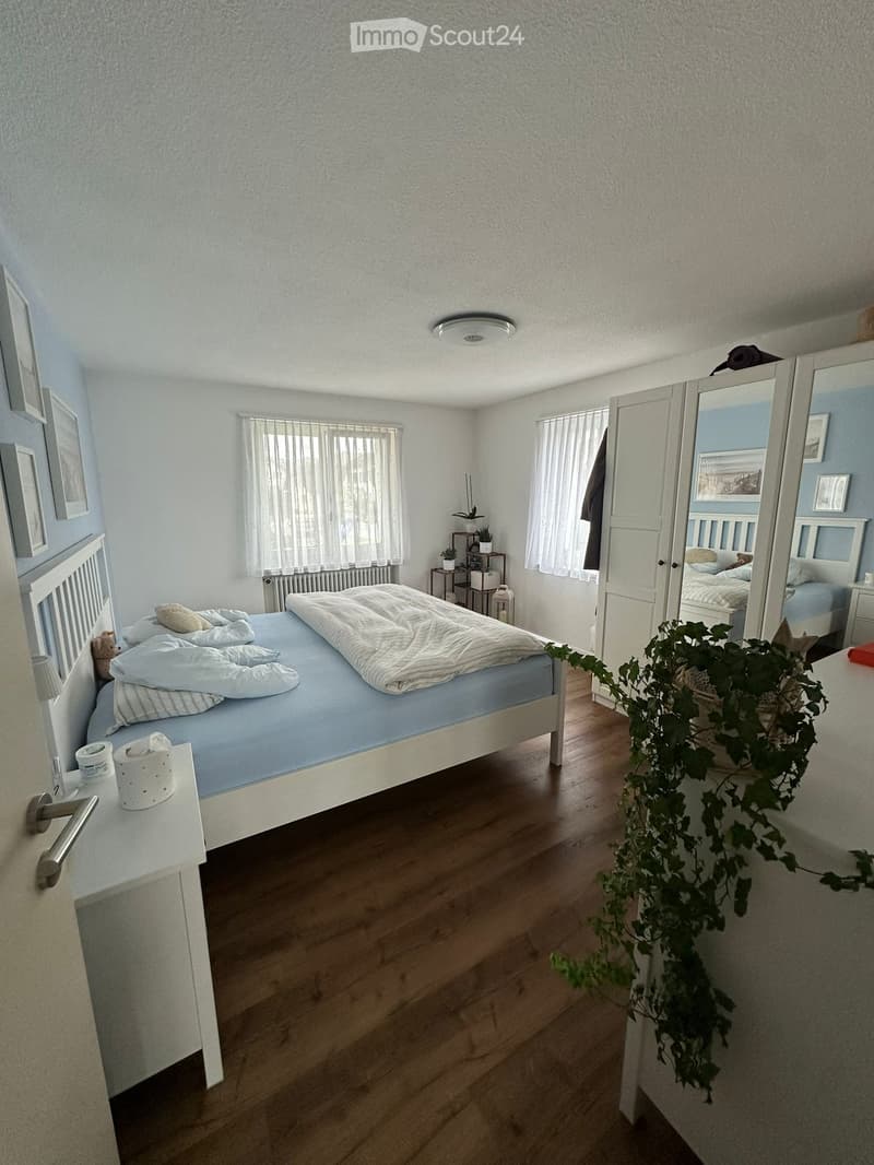 Cozy bedroom with a white bed, nightstand, mirror, wardrobe, and small plant. Light wooden flooring and white walls.