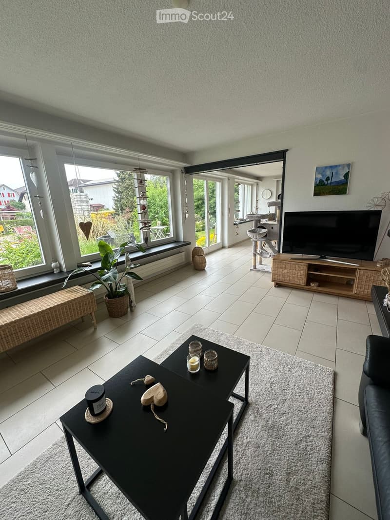 Bright living room with large windows, white tiled floor, grey sofa, black coffee table with candles, wicker bench, potted plant, and cat tree in the corner.