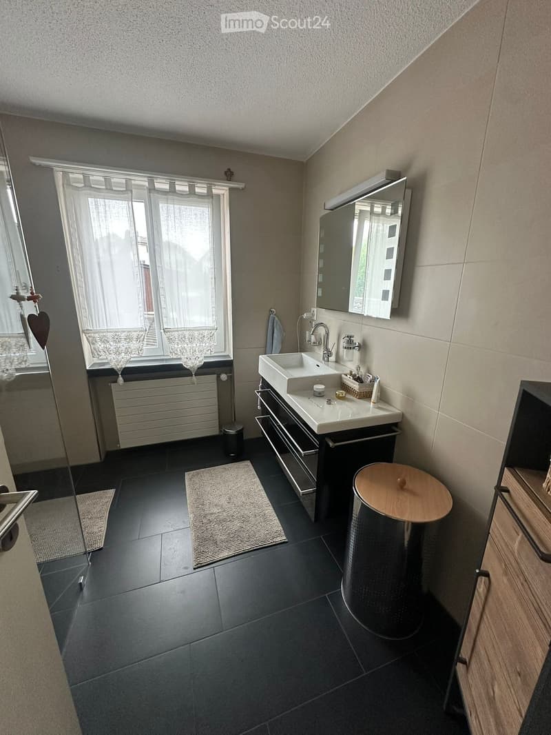 Modern bathroom with dark tiled floor, white sink, mirror, and a small wooden stool. Features a window with sheer curtains and a radiator below.