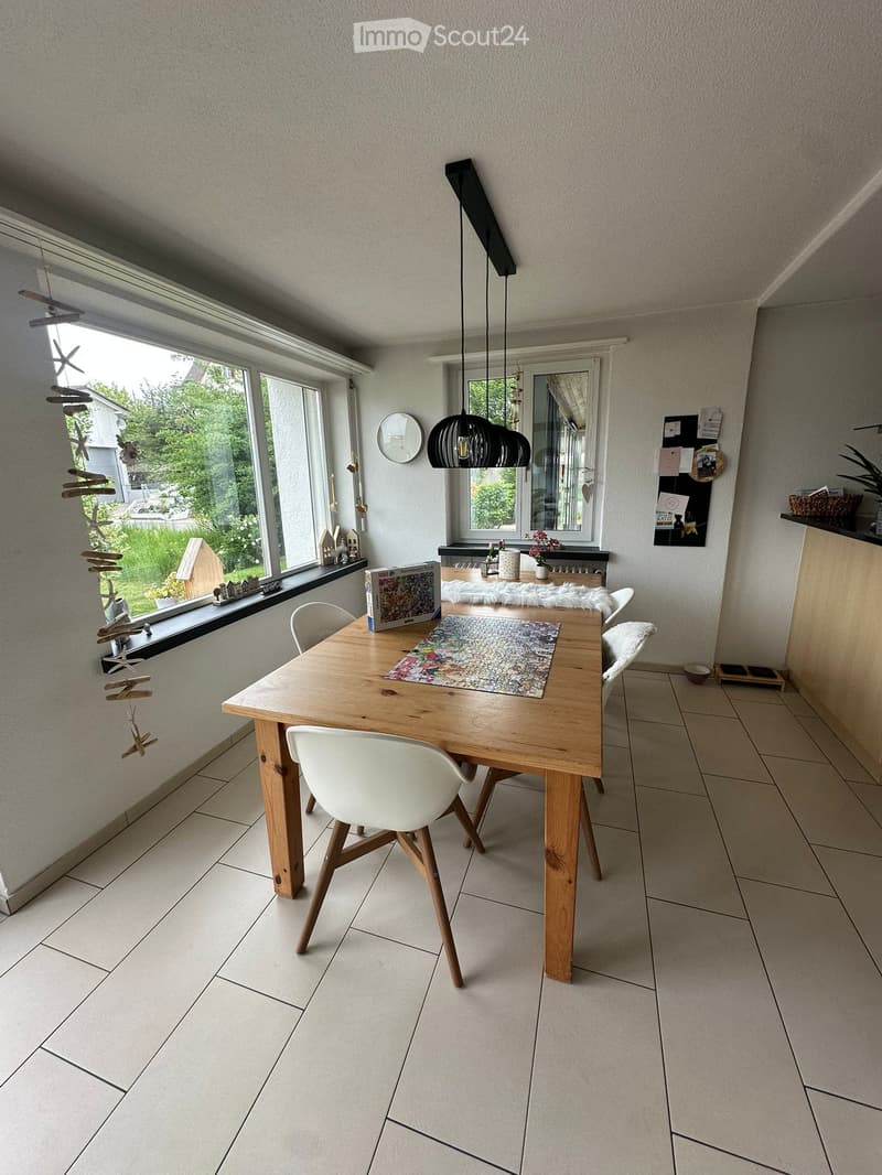 Bright dining area with a wooden table, white chairs, pendant light, and large windows overlooking a garden.