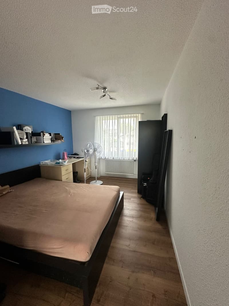 A furnished bedroom with a bed, desk, fan, shelf, radiator, and chair. The room features a wooden floor and a window with white curtains.