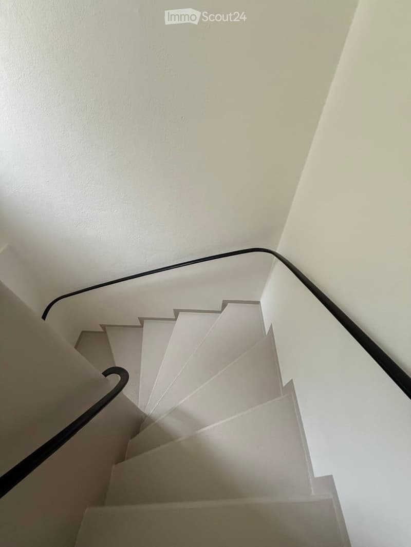 A modern, minimalist staircase with white steps and a sleek black handrail, set against clean white walls.