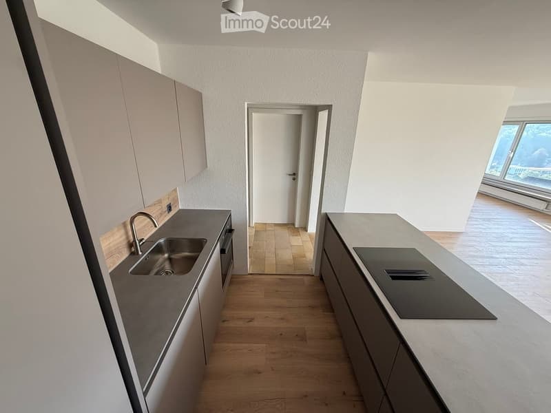Modern kitchen with light wooden flooring, white cabinets, and a dark countertop. Features a sink with a modern faucet and a built-in oven. A doorway leads to another room.