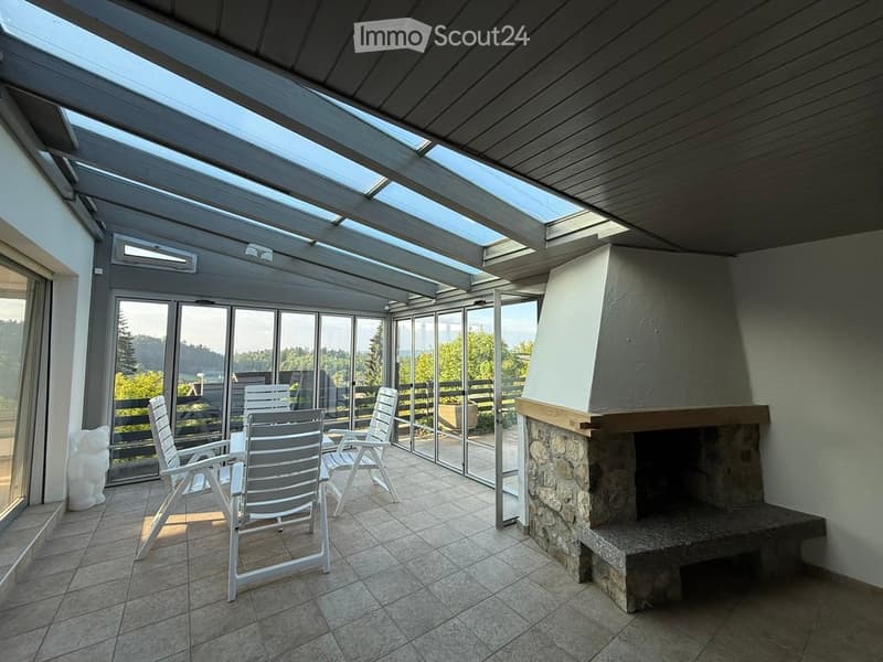 A spacious, covered balcony with a stone fireplace, tiled floor, and large glass windows offering a scenic view. The area includes a table and several white chairs.