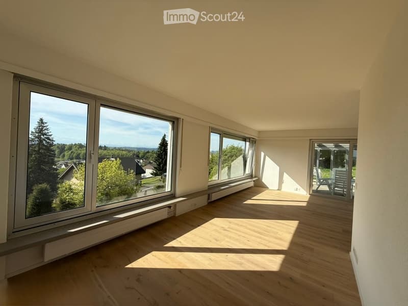 Empty room with wooden flooring, large windows, and a sliding glass door leading to a balcony area with outdoor furniture.