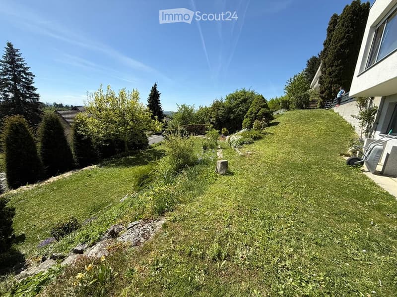 Well-maintained garden with green grass, shrubs, and trees, featuring a modern house with a balcony and a clear blue sky in the background.