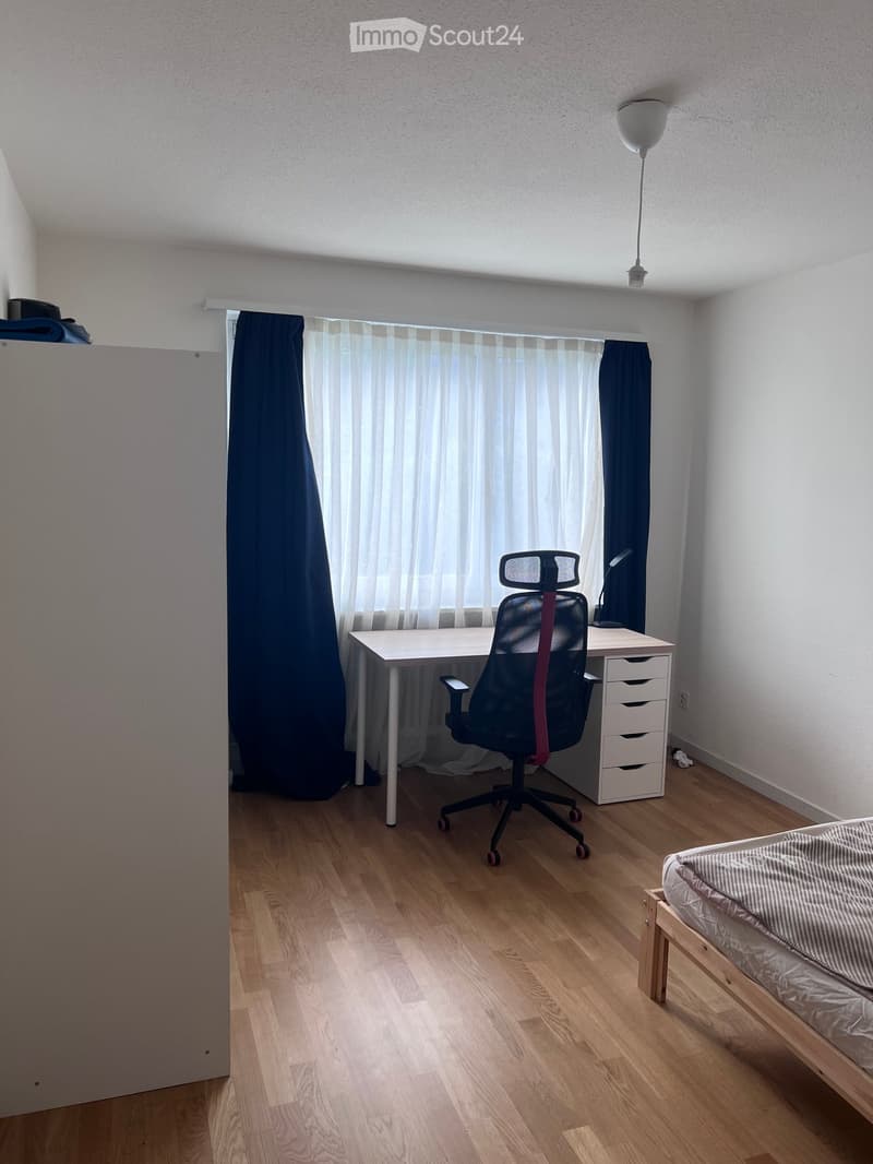 A small, well-lit room with a bed, desk, and chair. The room features wooden flooring, white walls, and a window with curtains.