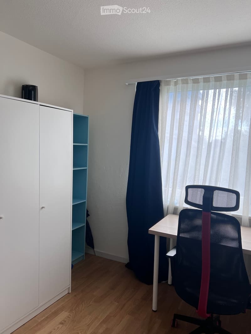 A small, furnished room with a wooden floor, white walls, a large window with blue and white curtains, a white cabinet, a small table, and a black office chair.