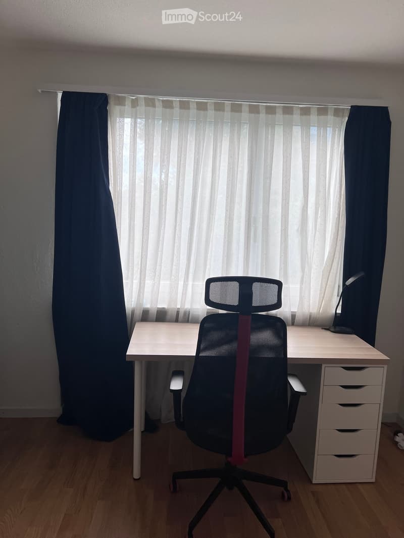A well-lit room with a wooden desk, ergonomic chair, lamp, and drawer unit, featuring large windows with sheer curtains and dark side curtains.