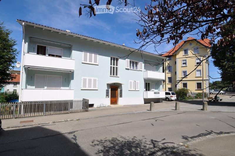 Two-story house with light blue exterior, white shutters, and balconies. Features a wooden front door and a small fence along the sidewalk.