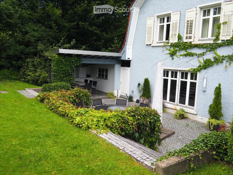A well-maintained garden with a paved pathway, green lawn, and a covered patio area with outdoor furniture.