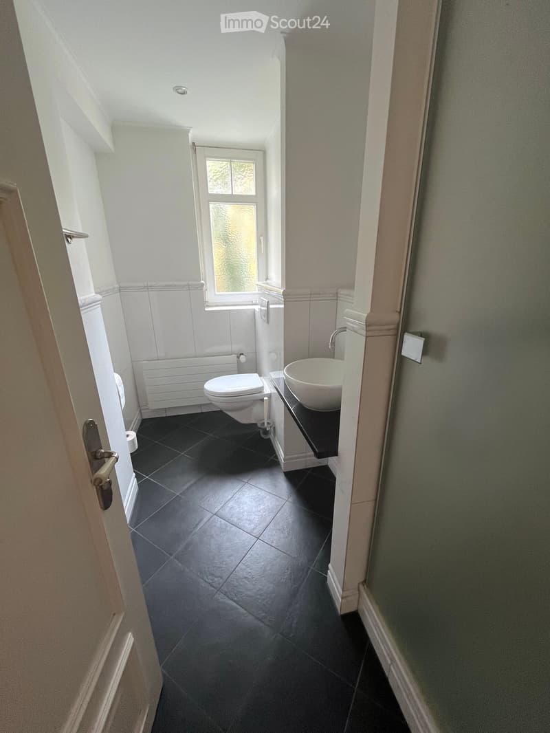 Modern bathroom with dark tiled floor, white toilet, white sink, and a window with frosted glass. The walls are painted in a light grey color with white trim around the window and door frame.