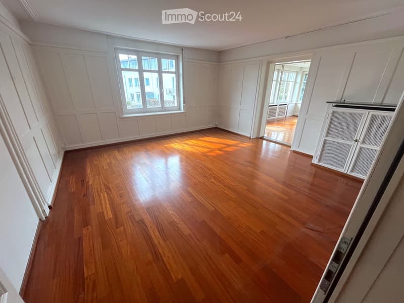 Empty room with wooden flooring, white walls, and large windows allowing natural light. The space is clean and unfurnished, with visible baseboards and radiators.