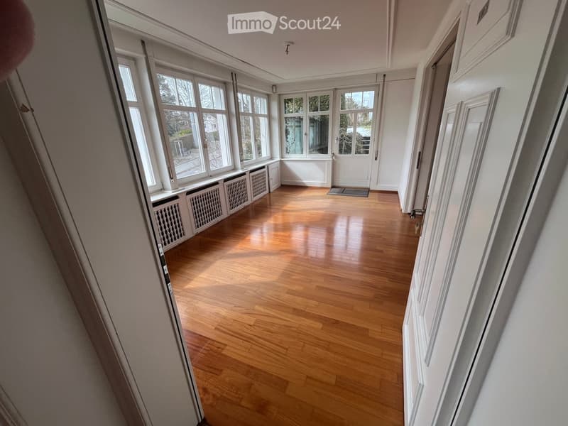 Empty hallway with wooden flooring, white walls, large windows, radiators, and doors leading to other rooms.