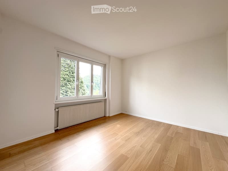 Empty room with light wooden flooring, white walls, and a large window with white frames. A radiator is visible beneath the window.