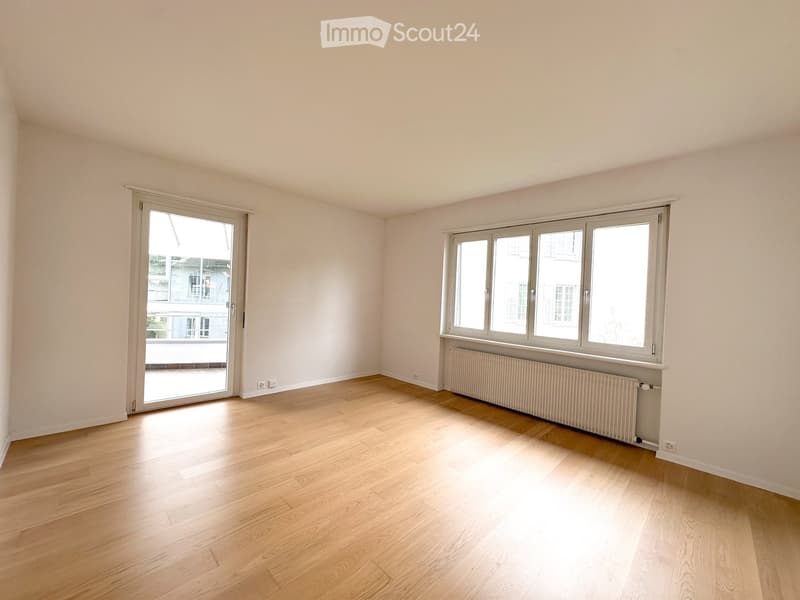 Empty room with light wooden flooring, white walls, large windows, and a sliding glass door leading to a balcony.