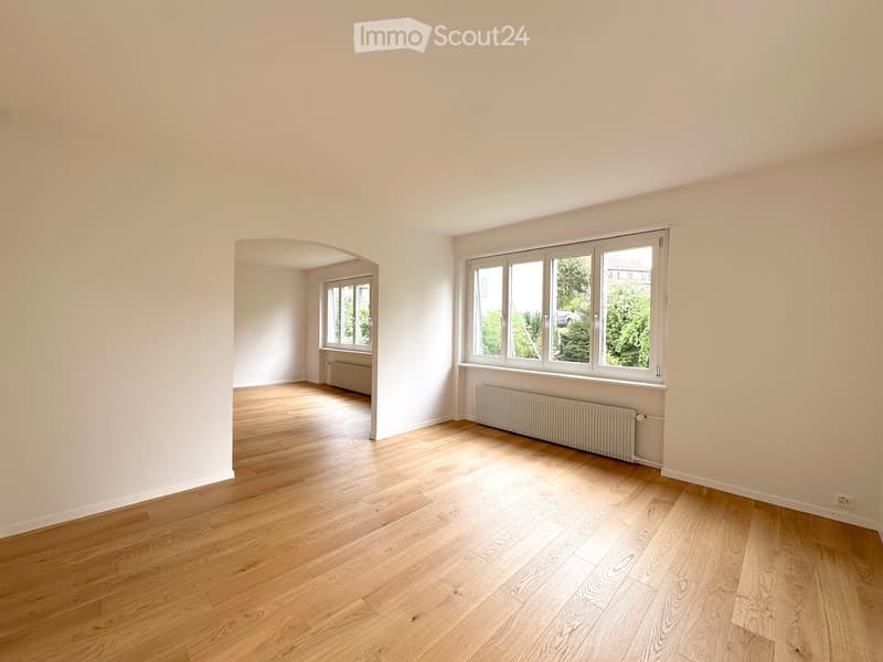 Empty room with light wooden flooring, white walls, and large windows allowing natural light. Radiators are visible under the windows.